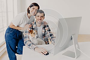 Indoor shot of happy family couple pose near computer monitor, work together remotely, have own business, earn money online, look