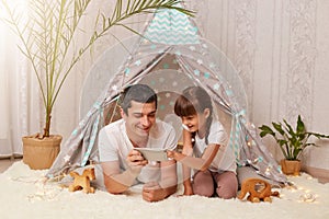 Indoor shot of happy family spending time at home, father with his daughter watching cartoons or funny video, expressing positive