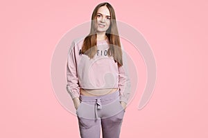Indoor shot of happy fit woman wears casual sportswear, keeps hands in pockets, stands alone against pink backgrpound, being in go