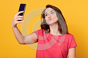 Indoor shot of happy flirting young girl taking pictures of herself through cell phone, charming female over yellow background,