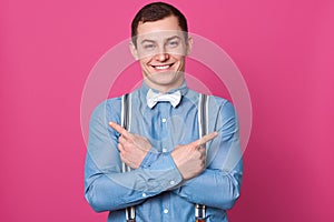 Indoor shot of happy funny young man laughing and looks excited, stands with crossing hands pointing fingers aside at copy space