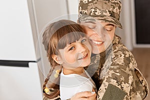 Indoor shot of happy military woman wearing camouflage uniform returning home after army, meeting her lovely daughter, hugging her