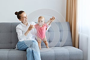 Indoor shot of happy satisfied woman with bun hairstyle wearing white shirt and jeans playing with her toddler daughter in pink