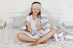 Indoor shot of happy smiling female with sleeping mask and being wrapped in blanket using cell phone, expressing positive emotions