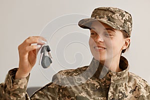 Indoor shot of happy smiling woman soldier wearing camouflage uniform and hat, posing at home, holding and looking at key in hands
