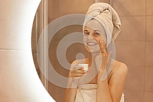 Indoor shot of happy smiling woman standing in front of mirror rubbing cosmetic cream on her face, putting on moisturizer on her