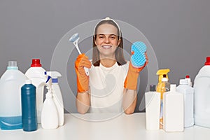 Indoor shot of happy smiling woman in white T-shirt and orange gloves, holding brush and sponger for cleaning house, being