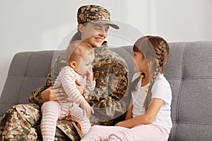 Indoor shot of happy soldier woman wearing camouflage uniform sitting on sofa with children, mother holding little daughter in