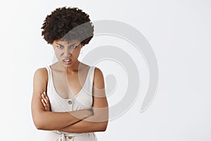 Indoor shot of hateful angry african american young woman with curly hair looking from under forehead with anger and