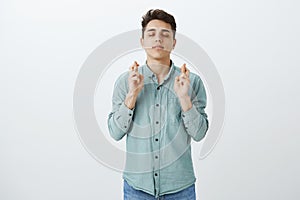 Indoor shot of hopeful confident young guy in shirt, closing eyes and smiling from happiness, crossing fingers while