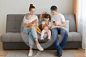 Indoor shot of husband with wife sitting on cough with their children and using cell phones, looking at device` s displays, gadge