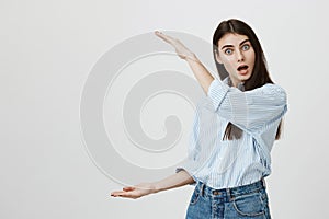 Indoor shot of impressed and amazed european woman with opened mouth and widened eyes shows something big with hands on