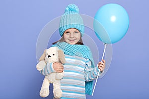 Indoor shot of little charming girl in winter hat with pom pom, scarf, and striped shirt, holding her favourite soft toy, stands