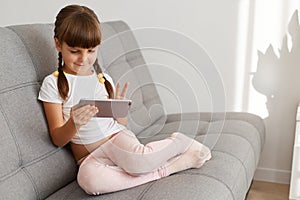 Indoor shot of little cute dark haired female child wearing white casual t shirt using cell phone sitting on couch, looking at