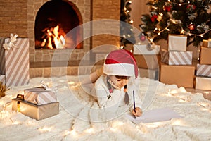 Indoor shot of little cute girl wearing white sweater and red hat, lying on floor on soft carpet in festive decorated room,