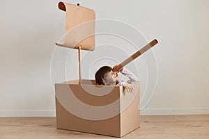 Indoor shot of little girl posing at home in a cardboard ship, play captains and sailors, holding spyglass and looking far,