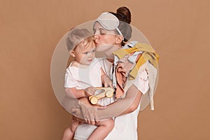 Indoor shot of loving mother standing with her infant daughter isolated over brown background, kissing her baby, expressing love