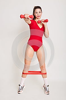 Indoor shot of magnetic beautiful fitness trainer holding dumbbells in both hands, using elastic band for fitness, wearing red
