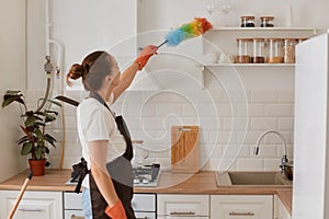 Indoor shot of maid wiping the dust with ppduster in the kitchen, housewife with bun hairstyle doing household chores, cleaning