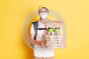 Indoor shot of man courier with thermo backpack in white T-shirt, cap and protective mask isolated over yellow background, holding