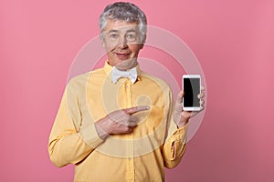Indoor shot of mature grey haired man in yellow shirt with bow tie, holding mobile phone in hands and pointing at black blank