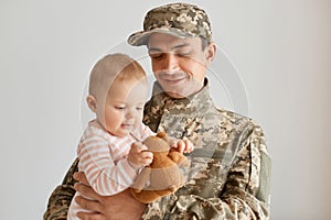 Indoor shot of military father came from the war or the exercises and cheerfully holding baby with soft toy, being happy to return