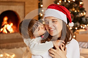 Indoor shot of mother and her little daughter hugging each other, having good mood, little cute girl kissing her mommy, merry