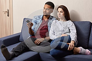 Indoor shot of multiethnic couple watch television at home on comfortable sofa. Black man holds remote control, switches on TV,