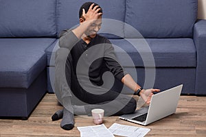 Indoor shot of overjoyed dark skinned man giggles as watches funny video on portable laptop computer, drinks coffee, surrounded