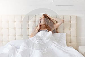 Indoor shot of peaceful attractive young lady relaxing in her bed in morning, yawning, covering her mouth with one hand, having