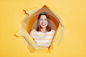 Indoor shot of playful young adult woman stands in torn paper hole, looking through breakthrough of yellow background and winking