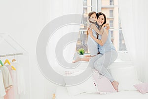 Indoor shot of pleasant looking female mother embraces her small kid, sit on window sill near bed, enjoy togetherness, have friend