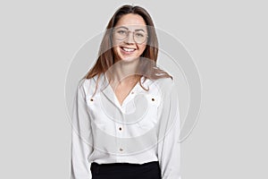 Indoor shot of pleasant looking smiling young female architect wears transparent round glasses, formal outfit, rejoices promotion