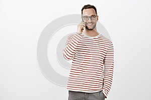 Indoor shot of pleased friendly adult office worker in striped pullover and glasses, talking on smartphone casually