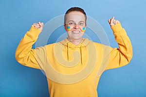 Indoor shot of positive happy Caucasian woman in yellow hoodie with Ukrainian flag on cheeks, waiting Ukraine victory, clenched