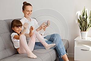 Indoor shot of positive satisfied woman wearing white shirt and jeans sitting on sofa with her child, people using phones, mother