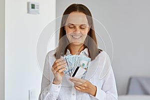 Indoor shot of positive young woman standing in light room at home, wearing white shirt, holding money and credit card, looking at