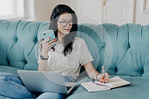 Indoor shot of pretty brunette female student watches webinar and makes notes in notepad, holds mobile phone, sits on comfortable