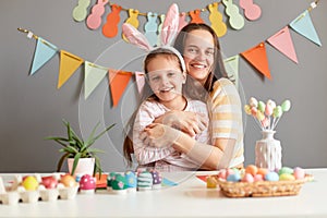 Indoor shot of pretty extremely happy mother and her daughter preparing for Easter, little child wearing bunny ears, woman hugging