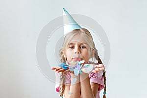 Indoor shot of pretty small kid with long pigtails, wears cone hat on head, blows colourful sheet of paper, looks into