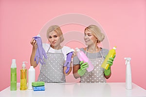 Indoor shot of pretty young blonde housewives dressed in basic t-shirts and aprons sitting over pink background with multi-colored