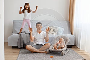 Indoor shot of relaxing handsome brunette father spensding time with his daughters while sitting on floor near sofa and practicing