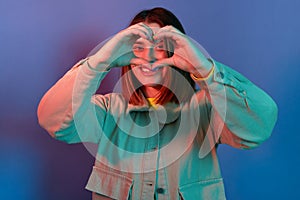 Indoor shot of romantic lovely woman wearing stylish jacket posing isolated on neon light background, making heart shape gesture