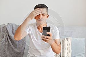 Indoor shot of sad upset man wearing casual style white t shirt sitting on sofa, using mobile phone with sorrow expression,