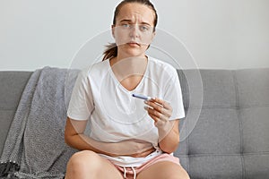 Indoor shot of sad young adult Caucasian woman holding pregnancy test in hands, touching her belly, looking at camera with sorrow