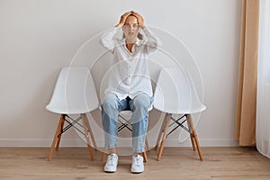 Indoor shot of scared Caucasian young adult woman with ponytail wearing white shirt and jeans, sitting on chair against light wall