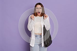 Indoor shot of scared frighted woman wearing stylish clothing holding bag posing isolated over purple background, standing with