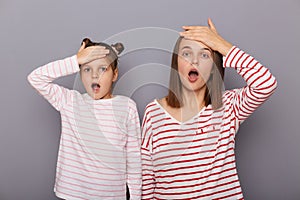 Indoor shot of scared shocked woman and little girl with hair buns wearing casual clothes standing isolated over gray background,