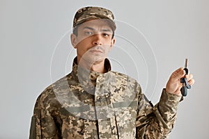 Indoor shot of serious Caucasian man soldier wearing camouflage uniform and cap, posing in his new apartment with keys in hands,