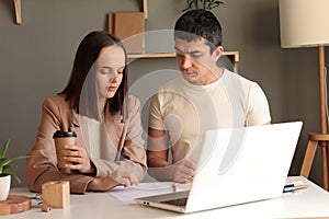 Indoor shot of serious concentrated man and woman sitting at table together in front of laptop working together and studying